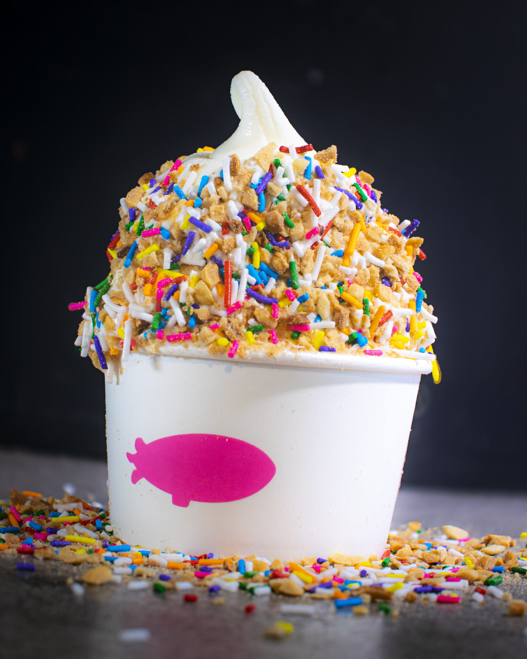 A vanilla soft serve ice cream in a white cup with Twinkle Nut Crunch Coat topping. The topping is all over the soft serve, and on the counter in an astheticlly pleasing and chaotic way