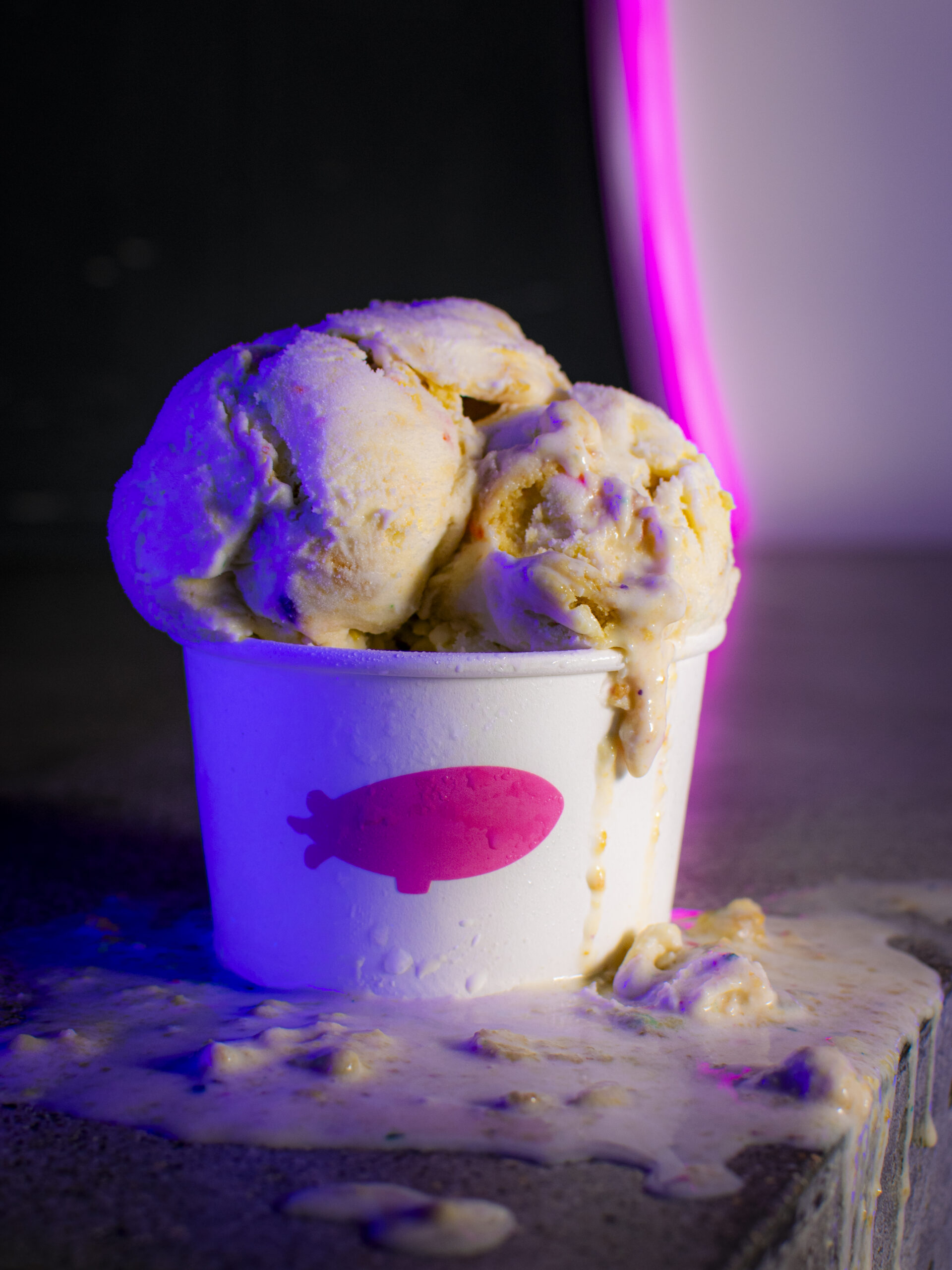A vanilla soft serve ice cream in a white cup with Twinkle Nut Crunch Coat topping. The topping is all over the soft serve, and on the counter in an astheticlly pleasing and chaotic way