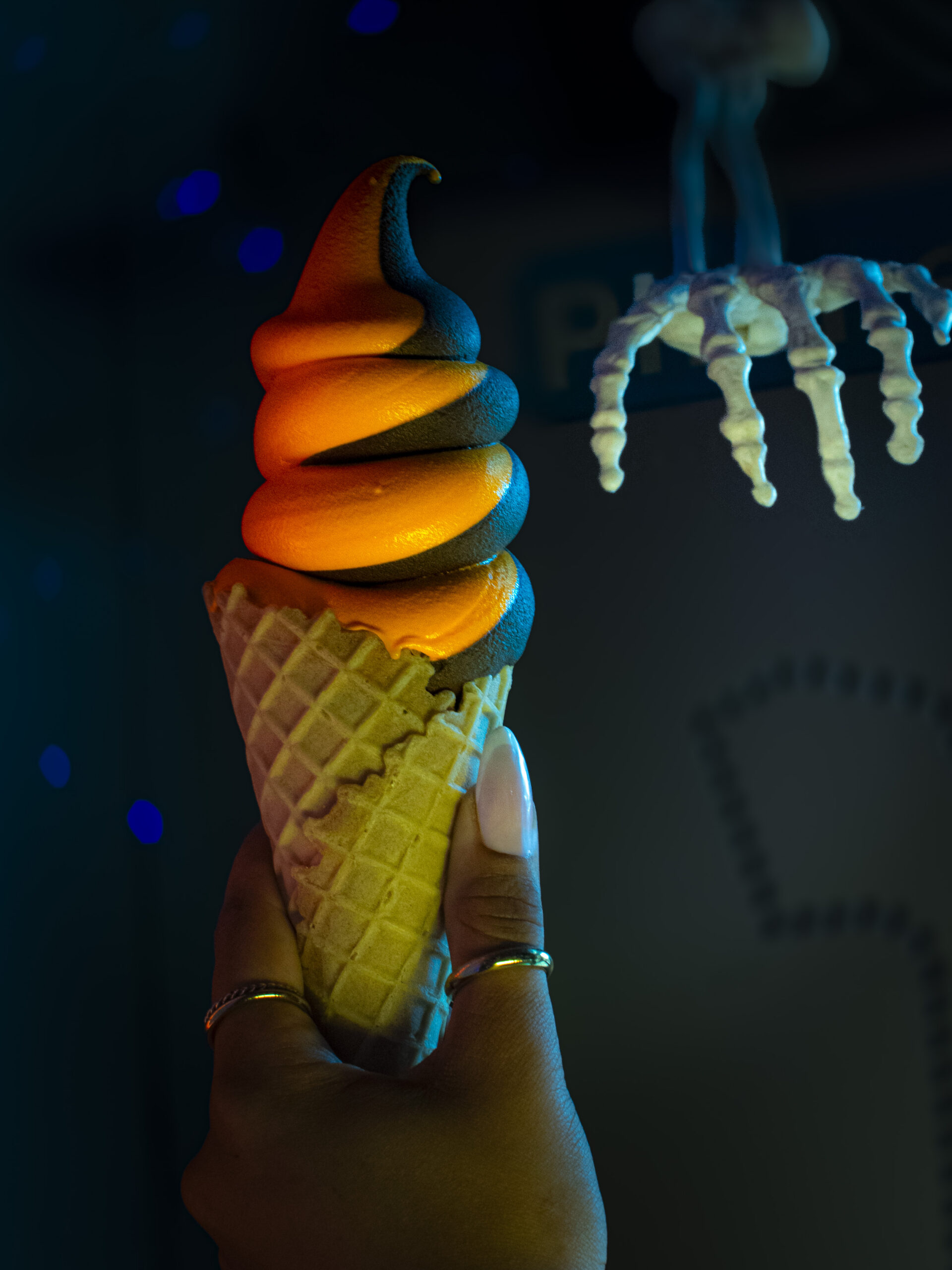 A woman's hand holds up a Halloween-themed orange and black soft serve ice cream in a waffle cone. There is a skeleton hand reaching towards the cone in the background.